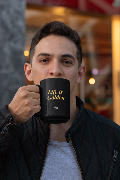 A man holding a black ceramic mug with the phrase 'Life is Golden' printed in gold lettering, set against a warm, softly blurred storefront background. 