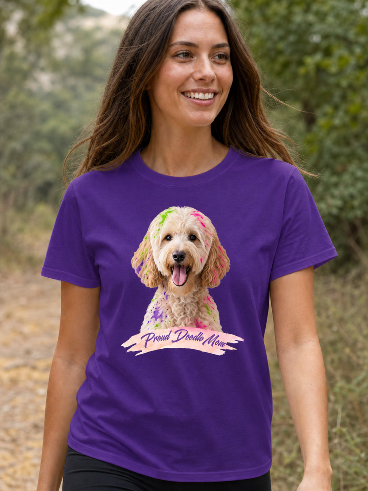 A woman wearing a purple short-sleeve crewneck t-shirt featuring a graphic print of a goldendoodle dog with colorful paint on its fur and a banner reading 'Proud Doodle Mom'.