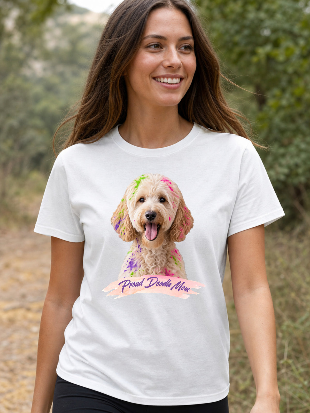 A woman wearing a white short-sleeve crewneck t-shirt featuring a graphic print of a goldendoodle dog with colorful paint on its fur and a banner reading 'Proud Doodle Mom'.