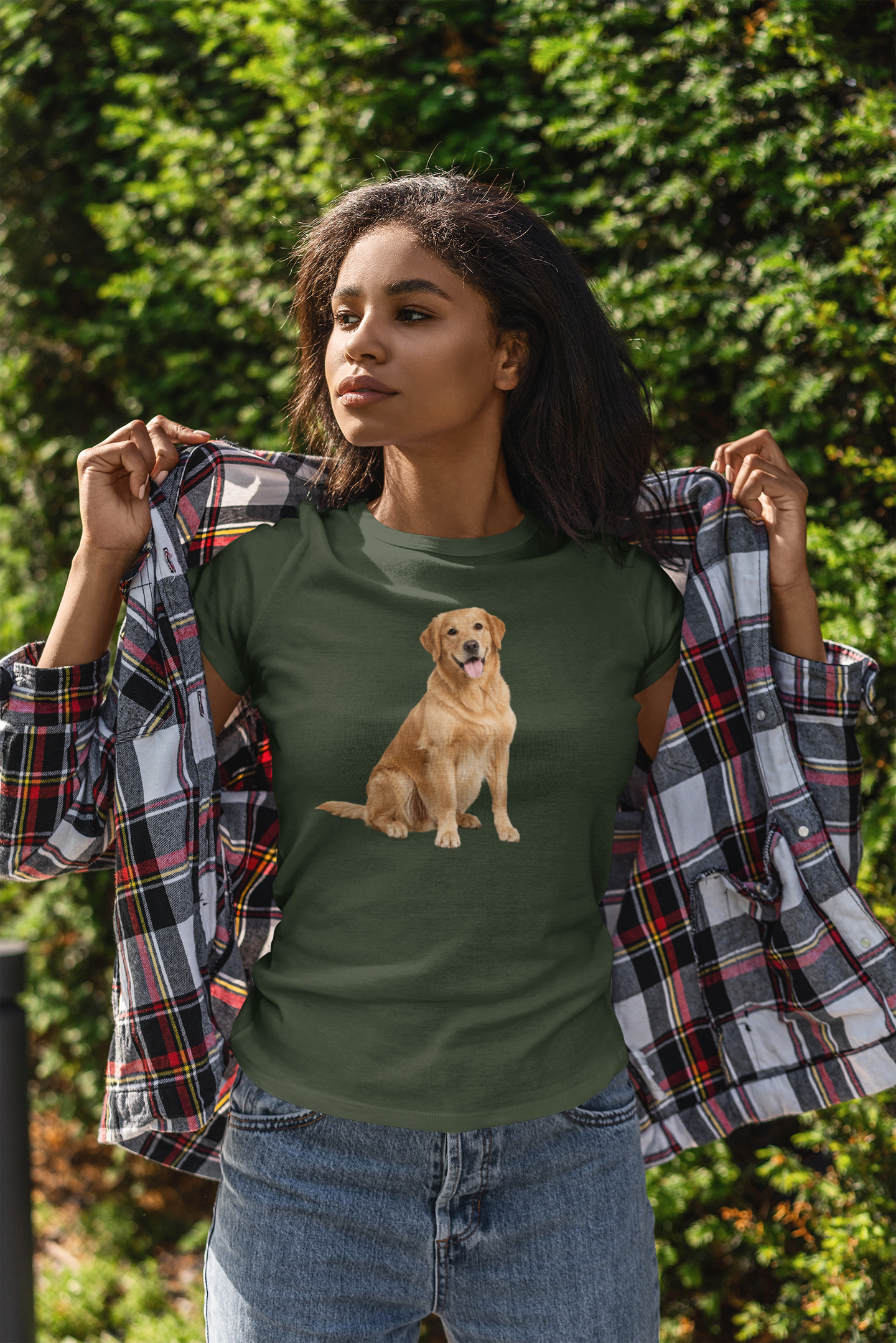 A woman outdoors wearing a forest green t-shirt with a graphic of a golden retriever dog, holding a black, red, and white plaid flannel shirt open over her shoulders. She is wearing blue jeans, with a background of green foliage.