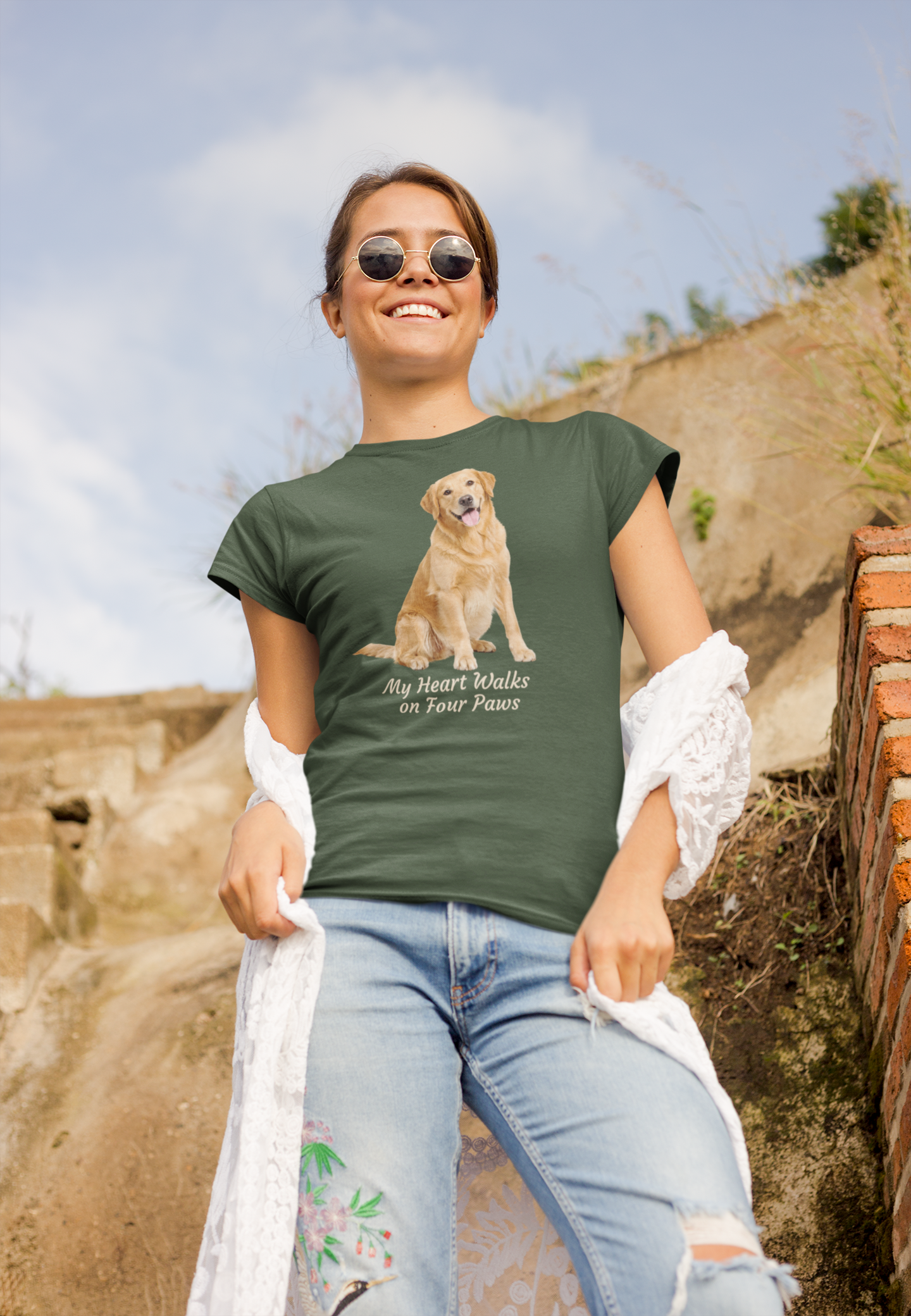 A woman wearing a forest green graphic t-shirt with a Golden Retriever print and the text 'My Heart Walks on Four Paws', paired with light blue jeans and sunglasses, standing outdoors against a rustic stone wall.