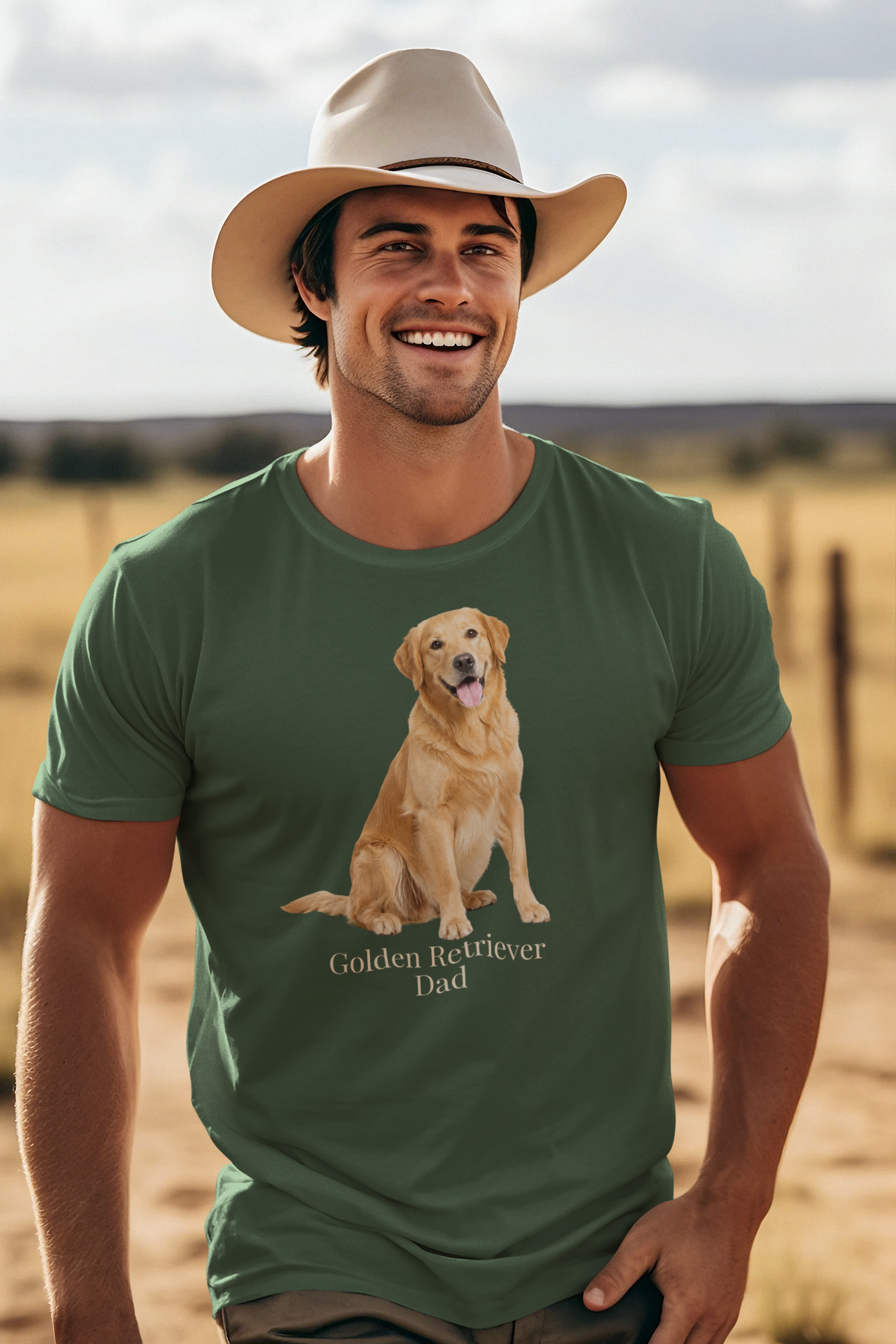 A smiling man wearing a beige cowboy hat and a forest green short-sleeve t-shirt with a graphic of a golden retriever dog sitting above the text 'Golden Retriever Dad' in beige font, set against a blurred outdoor ranch landscape.