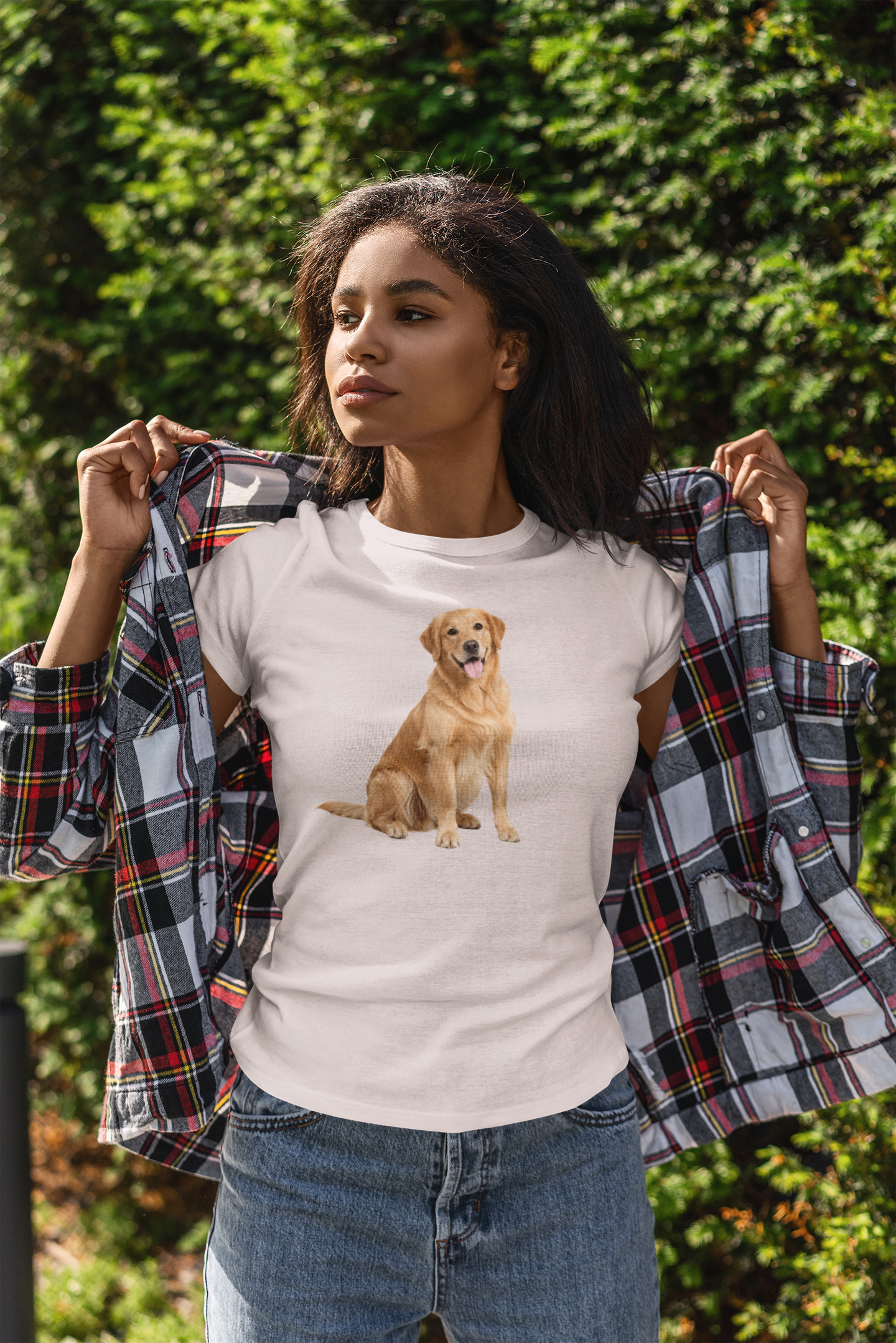A woman outdoors wearing an ice gray t-shirt with a graphic of a golden retriever dog, holding open a plaid flannel shirt over her shoulders. She is wearing blue jeans, with a background of green foliage.