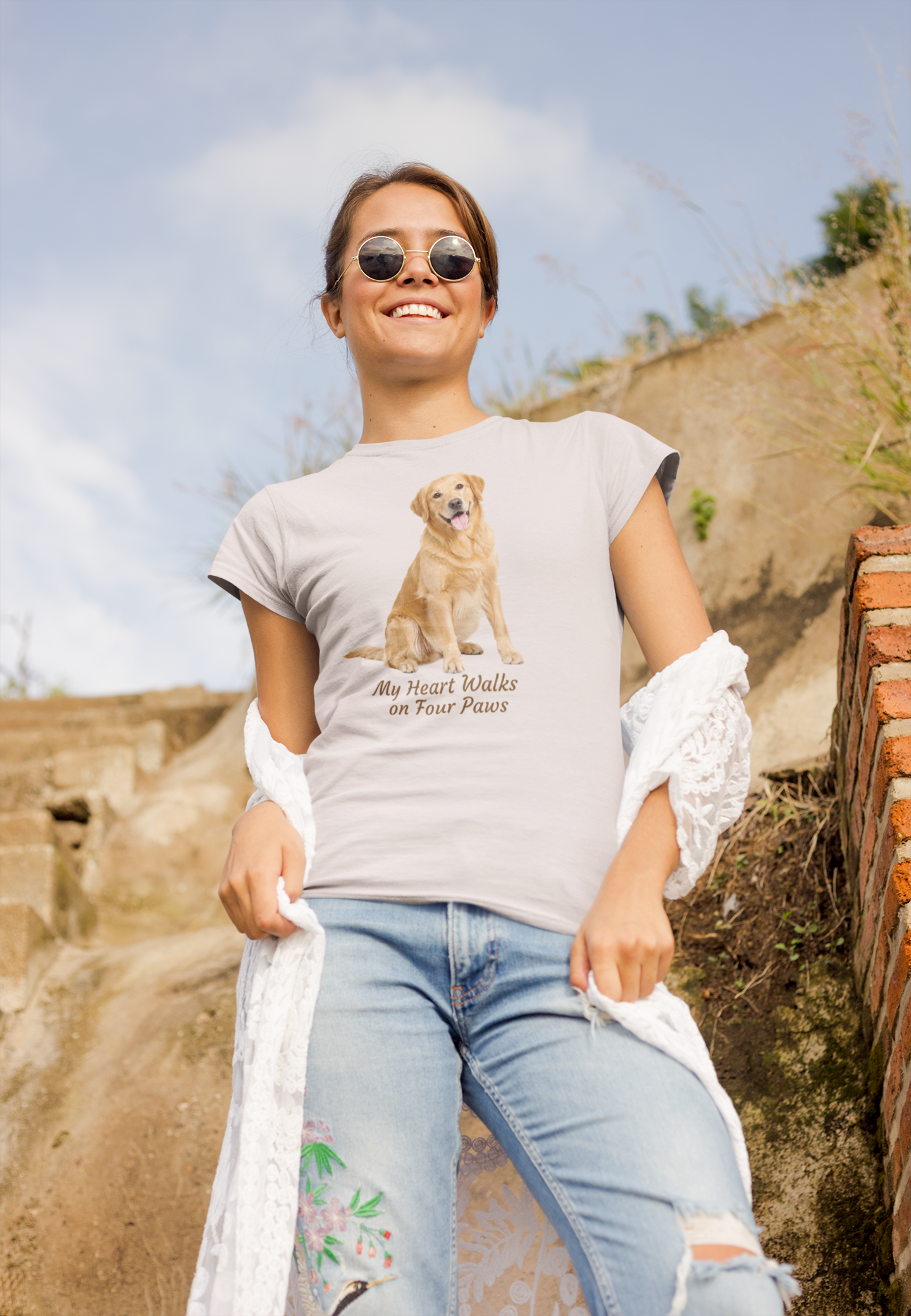 A woman wearing an ice gray graphic t-shirt with a Golden Retriever print and the text 'My Heart Walks on Four Paws', paired with light blue jeans and sunglasses, standing outdoors against a rustic stone wall.