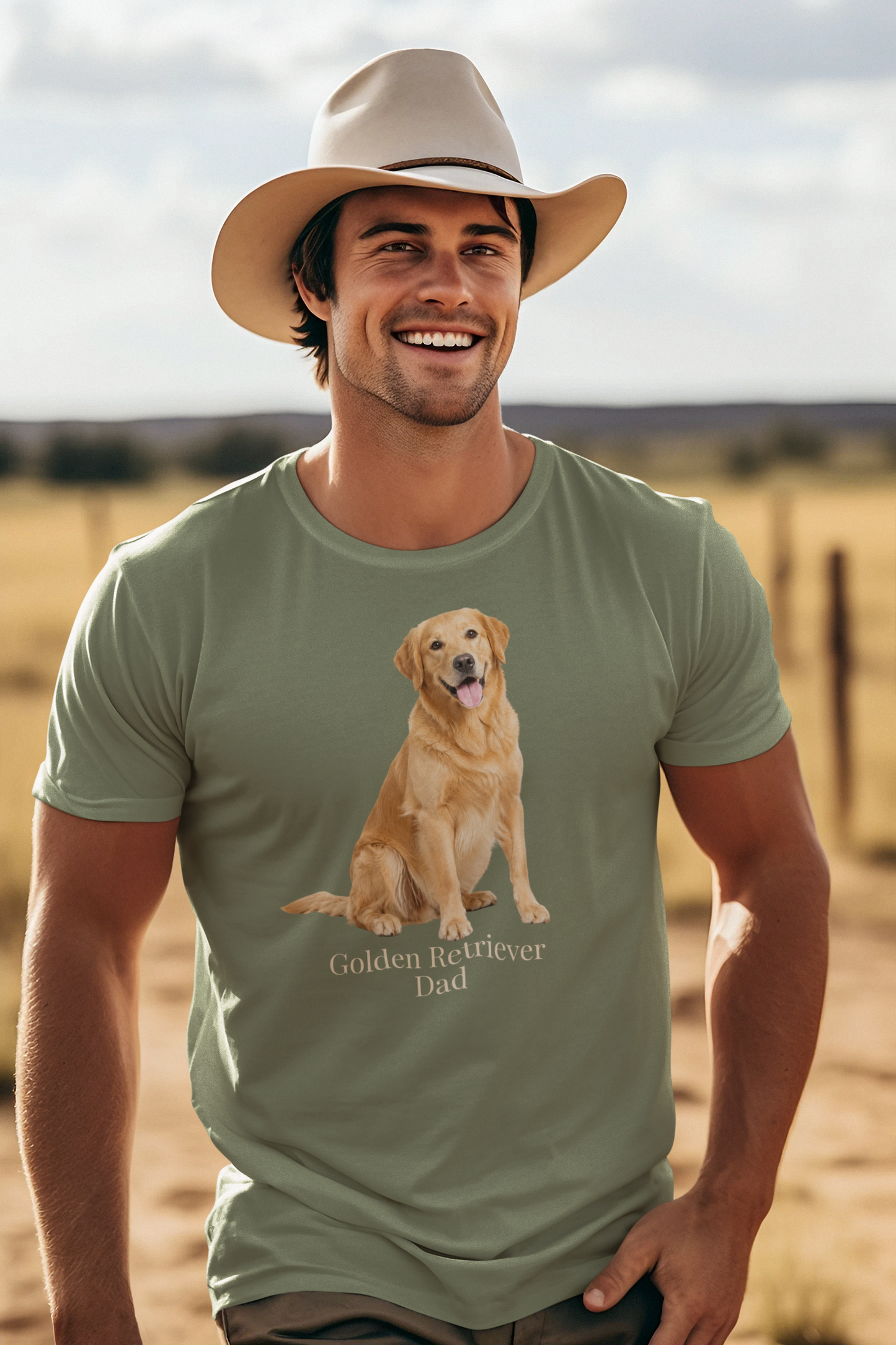 A smiling man wearing a beige cowboy hat and a military green short-sleeve t-shirt with a graphic of a golden retriever dog sitting above the text 'Golden Retriever Dad' in beige font, set against a blurred outdoor ranch landscape.