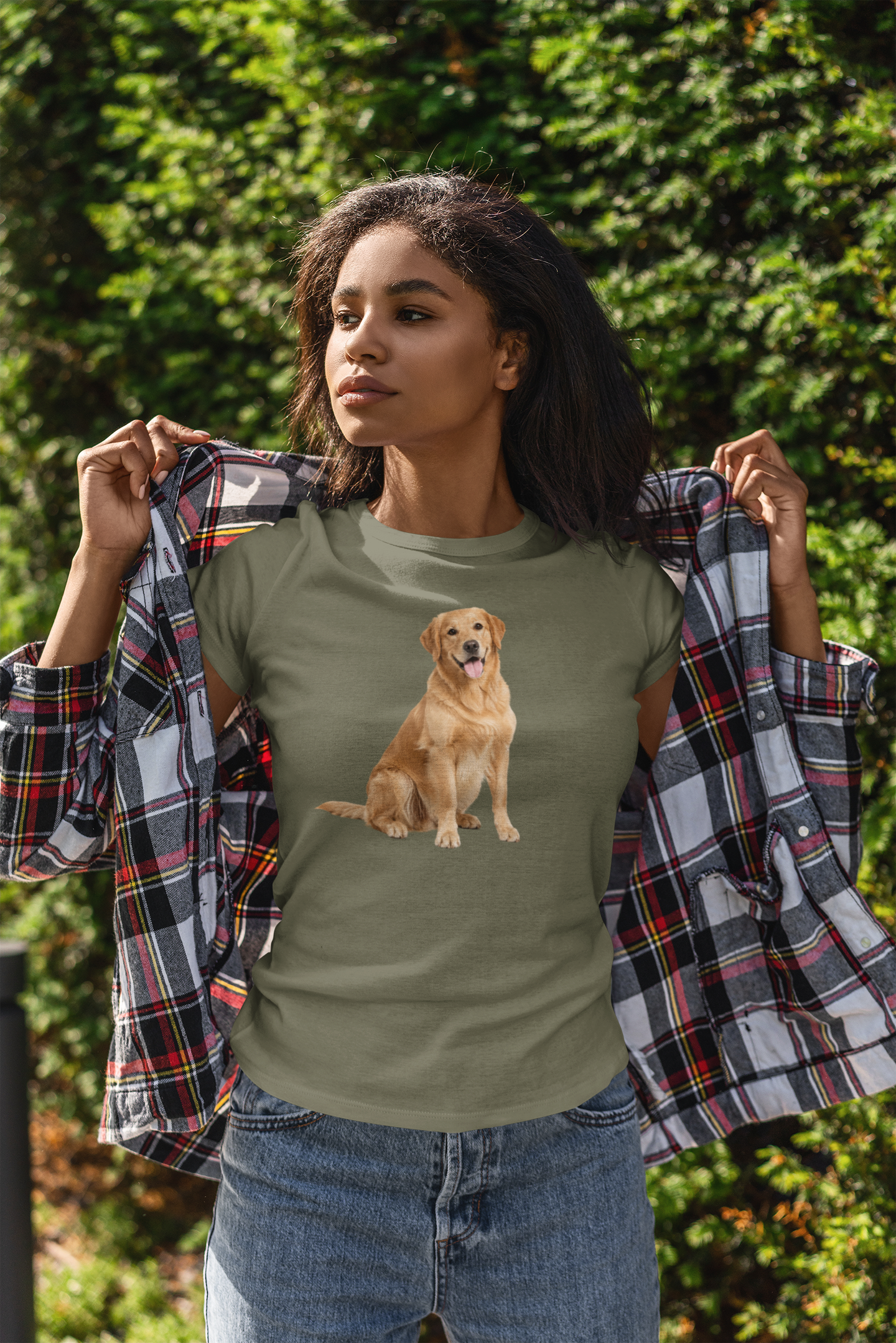 A woman outdoors wearing a military green t-shirt with a graphic of a golden retriever dog, holding open a plaid flannel shirt over her shoulders. She is wearing blue jeans, with a background of green foliage.