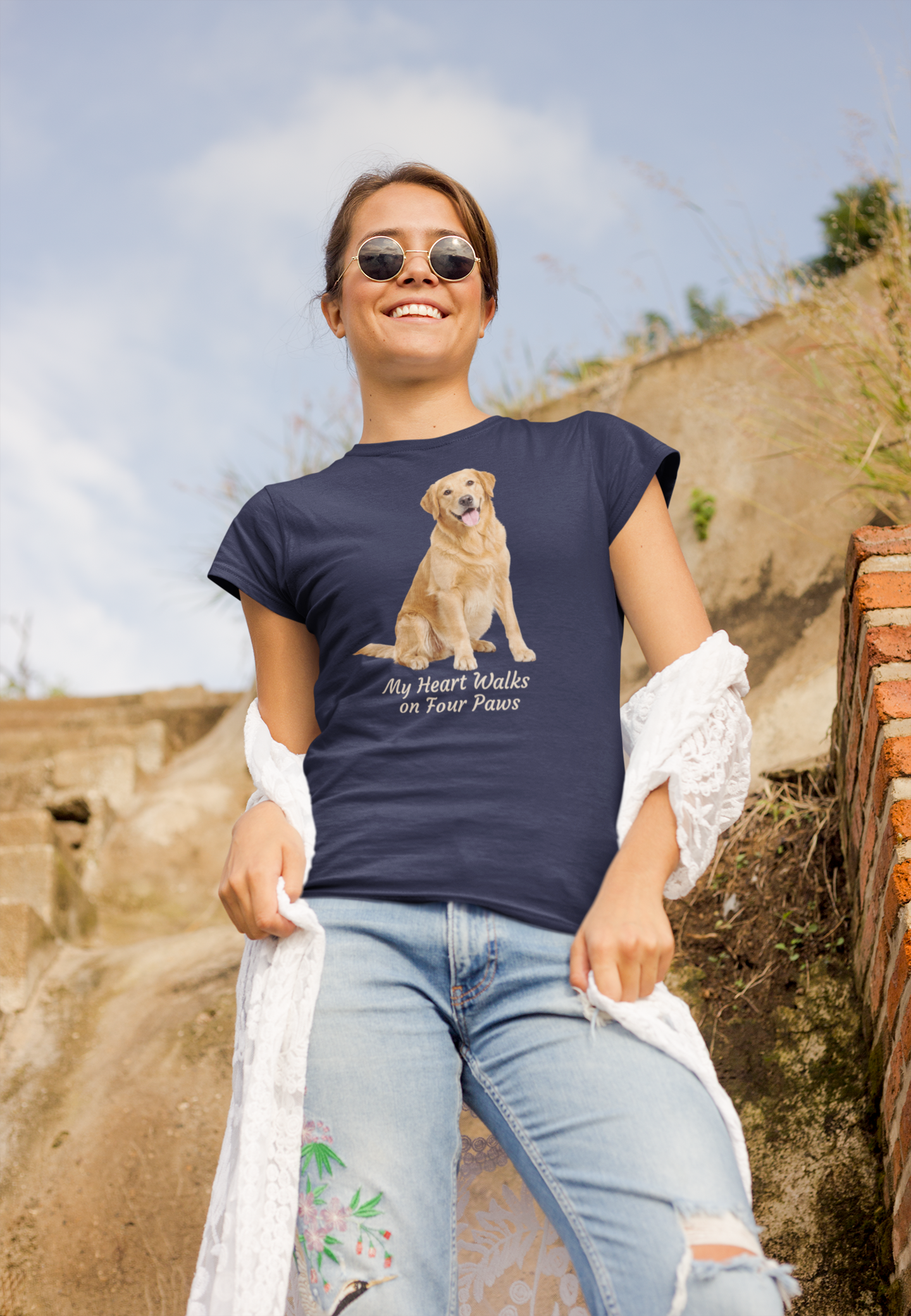 A woman wearing a navy graphic t-shirt with a Golden Retriever print and the text 'My Heart Walks on Four Paws', paired with light blue jeans and sunglasses, standing outdoors against a rustic stone wall.
