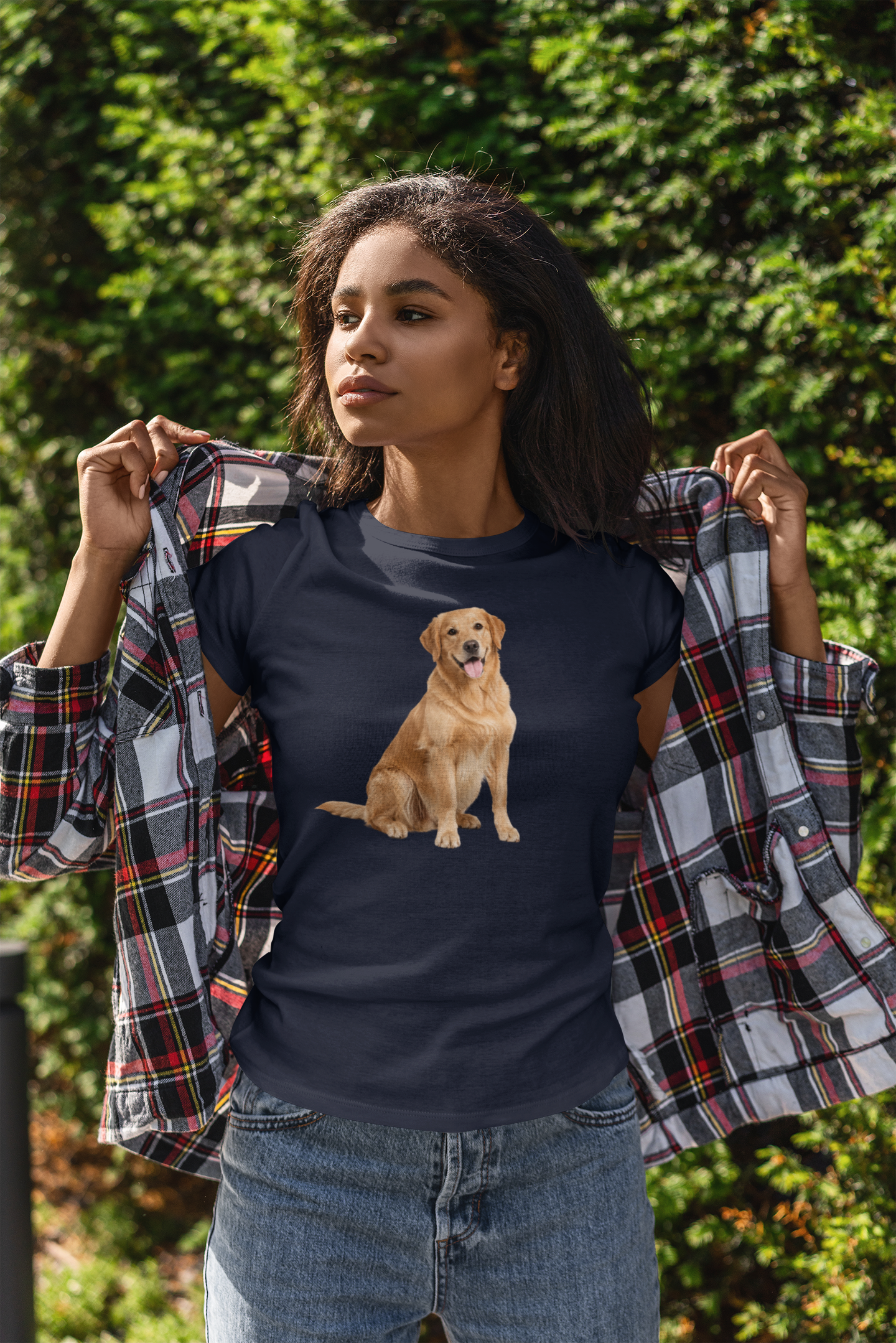 A woman outdoors wearing a navy blue t-shirt with a graphic of a golden retriever dog, holding open a plaid flannel shirt over her shoulders. She is wearing blue jeans, with a background of green foliage.