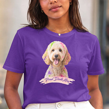 A woman wearing a purple short-sleeve crewneck t-shirt featuring a graphic print of a goldendoodle dog with colorful paint on its fur and a banner reading 'Proud Doodle Mom', paired with white pants.