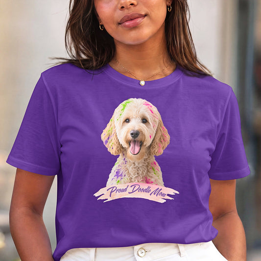A woman wearing a purple short-sleeve crewneck t-shirt featuring a graphic print of a goldendoodle dog with colorful paint on its fur and a banner reading 'Proud Doodle Mom', paired with white pants.