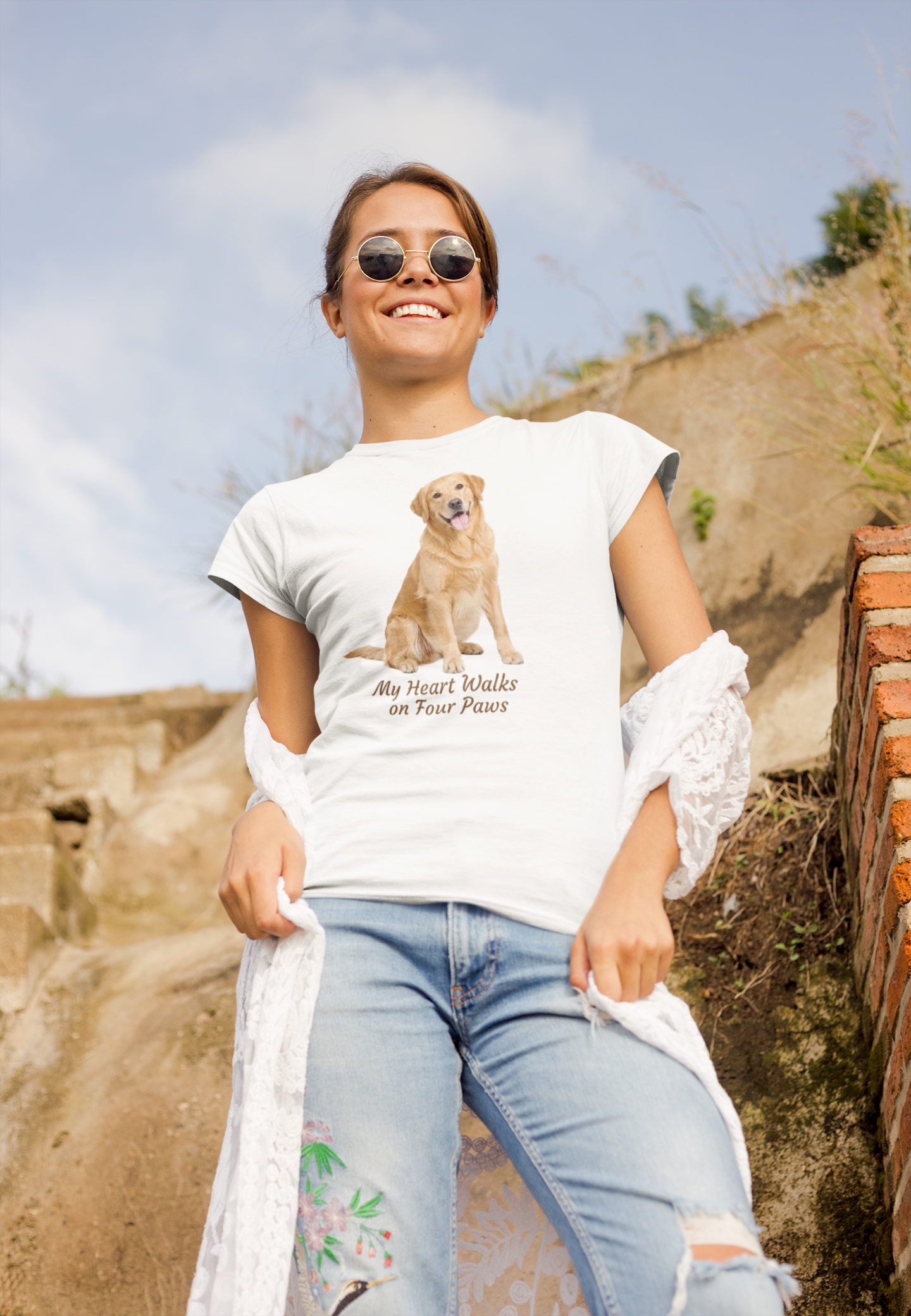 A woman wearing a white graphic t-shirt with a Golden Retriever print and the text 'My Heart Walks on Four Paws', paired with light blue jeans and sunglasses, standing outdoors against a rustic stone wall.