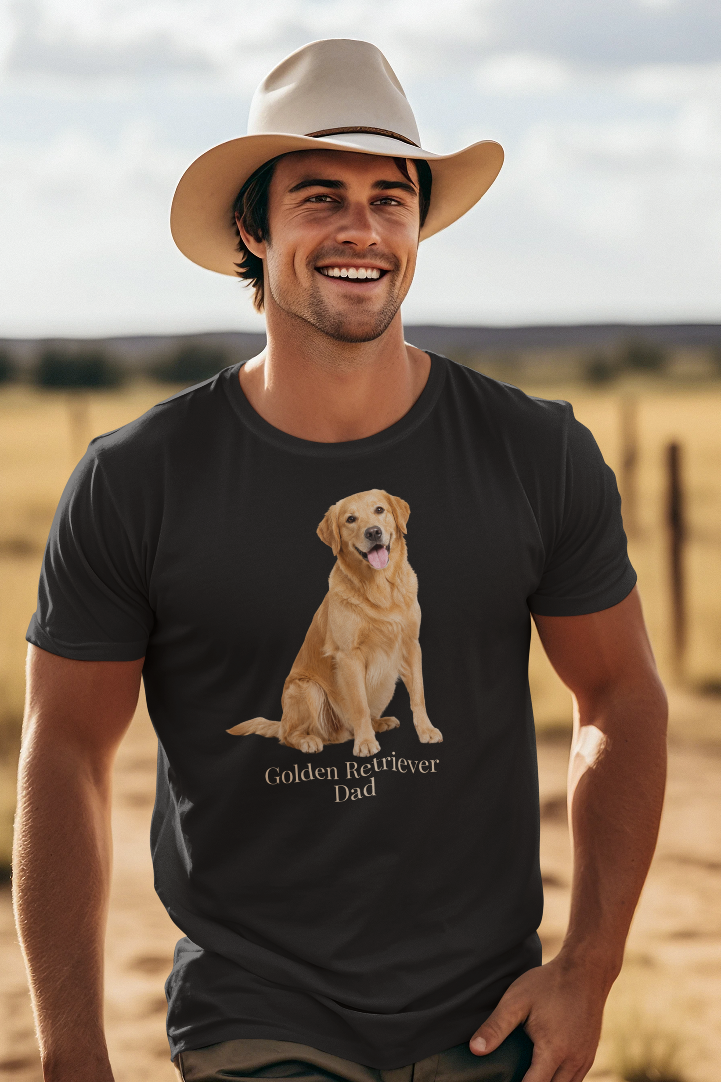 A smiling man wearing a beige cowboy hat and a black short-sleeve t-shirt with a graphic of a golden retriever dog sitting above the text 'Golden Retriever Dad' in beige font, set against a blurred outdoor ranch landscape.