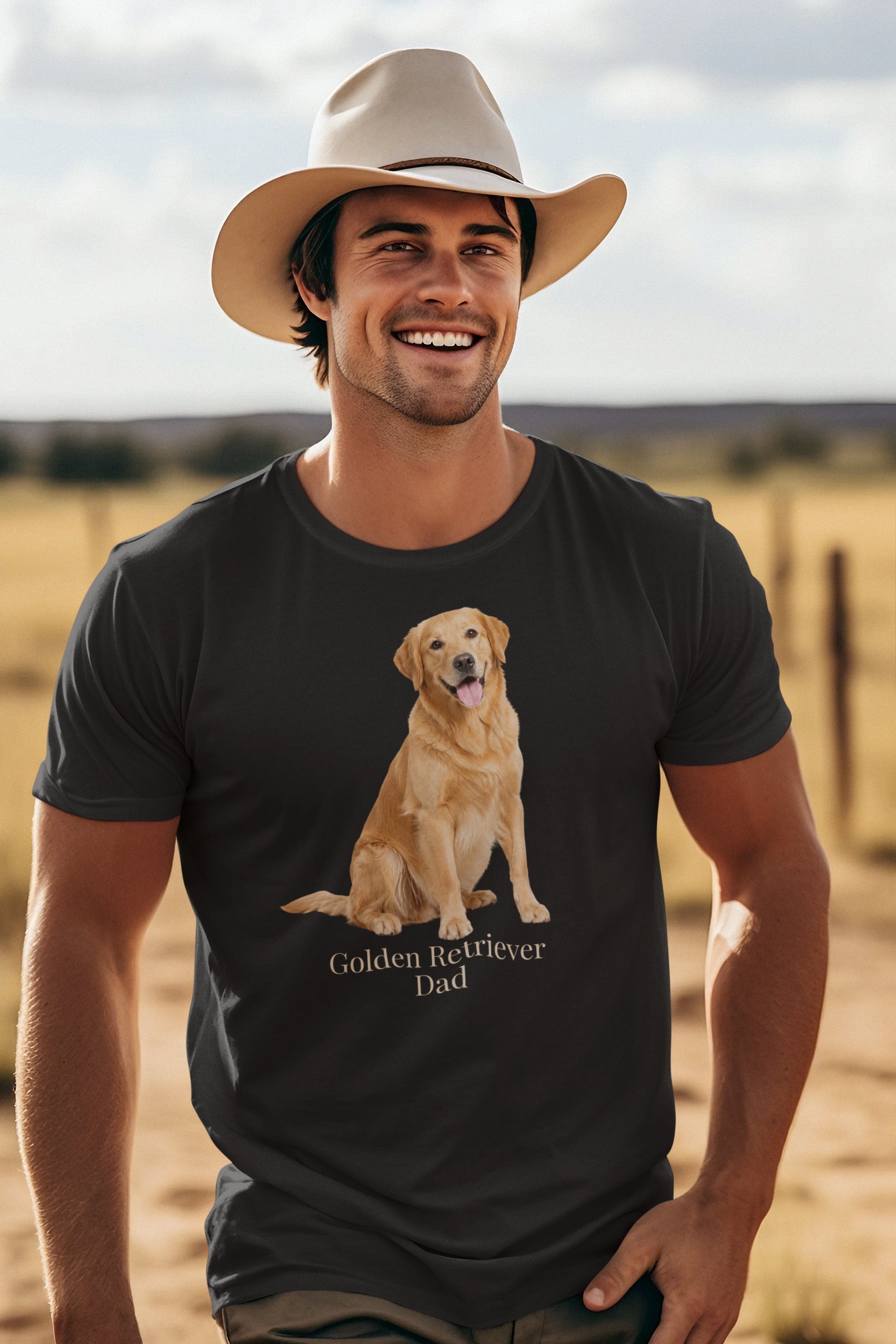 A smiling man wearing a beige cowboy hat and a black short-sleeve t-shirt with a graphic of a golden retriever dog sitting above the text 'Golden Retriever Dad' in beige font, set against a blurred outdoor ranch landscape.