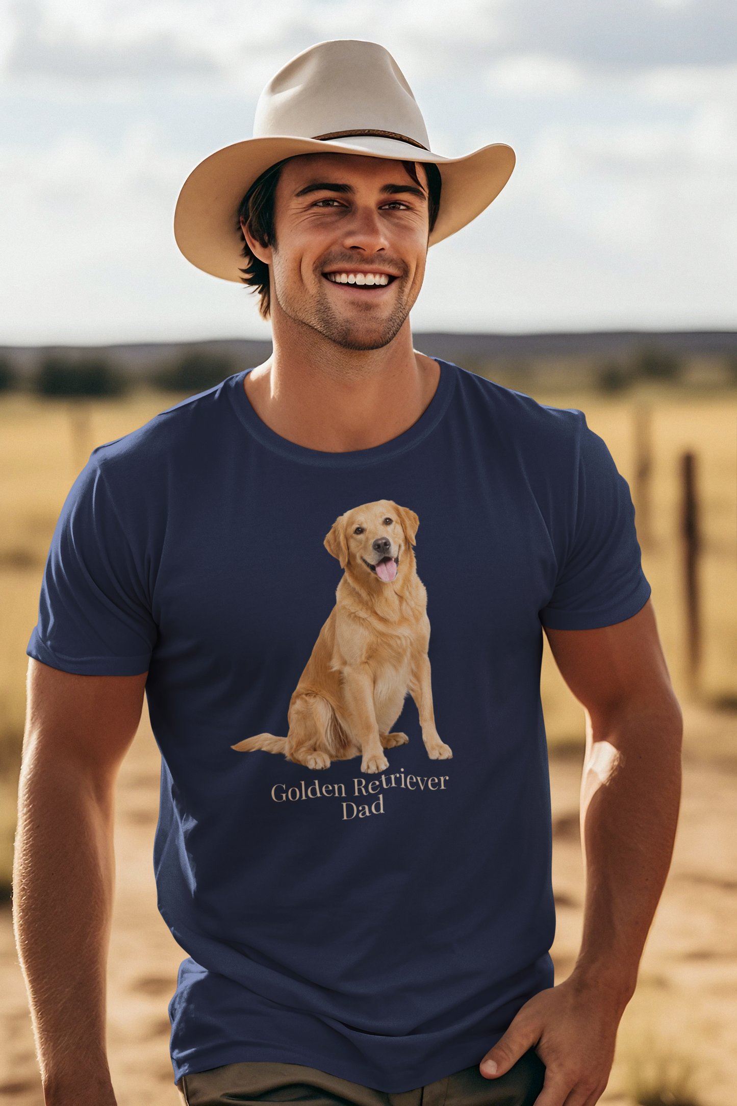 A smiling man wearing a beige cowboy hat and a navy blue short-sleeve t-shirt with a graphic of a golden retriever dog sitting above the text 'Golden Retriever Dad' in beige font, set against a blurred outdoor ranch landscape.