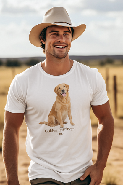 A smiling man wearing a beige cowboy hat and a white short-sleeve t-shirt with a graphic of a golden retriever dog sitting above the text 'Golden Retriever Dad' in dark brown font, set against a blurred outdoor ranch landscape.